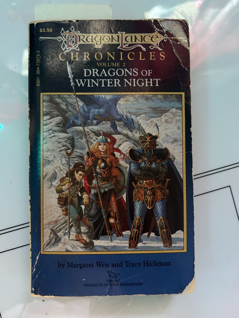 a photograph of an OLD copy of Dragons of Winter Night by Margaret Weis and Tracy Hickman. The cover shows three adventurers standing in the snow, and a blue dragon on the mountain behind them.