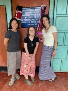 three women standing in front of a placard at a rural school in Kenya